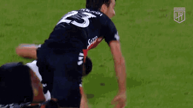 a soccer player in a black jersey with the number 23 on it is kicking the ball while another player is laying on the ground .