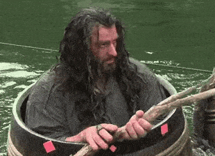 a man with long hair is sitting in a wooden barrel in the water holding a stick .