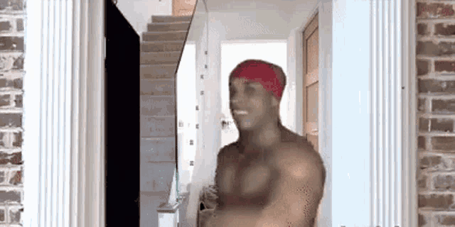 a shirtless man wearing a red bandana is standing in a hallway next to a staircase .