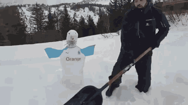 a man is shoveling snow next to a snowman with an orange shirt on it