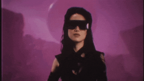 a woman wearing sunglasses and a black dress is standing in front of a pink background .
