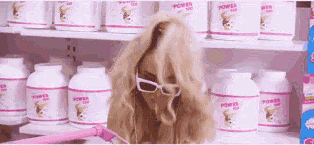 a woman wearing glasses is standing in front of a shelf of power 100 powder