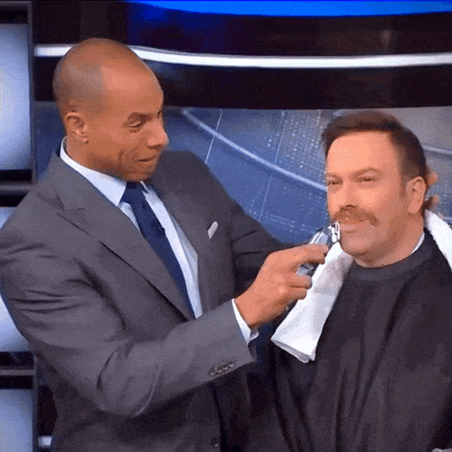 a man in a suit and tie is shaving another man 's mustache
