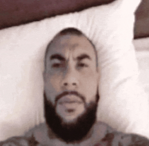 a man with a beard is laying in bed with a pillow on his head .