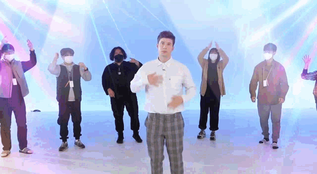 a man in a white shirt and plaid pants is standing in front of a group of people wearing face masks .