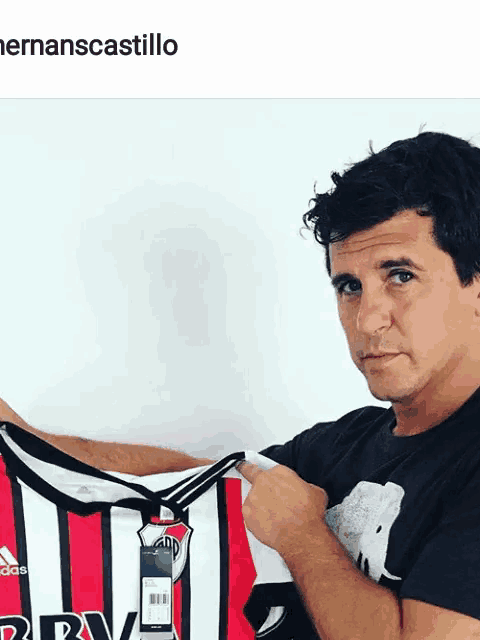 a man in a black shirt holds up a red and white adidas jersey