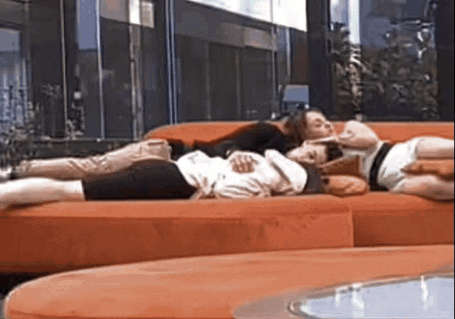 two people are laying on an orange couch in front of a large window