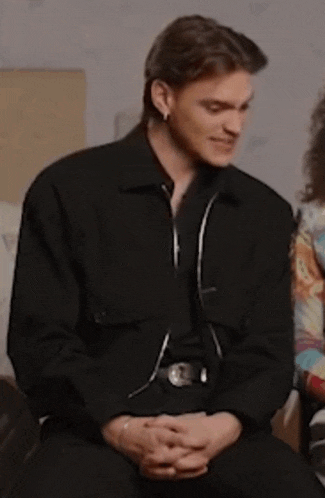 a man in a black jacket is sitting in a chair with his hands folded and smiling .