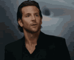 a man in a black suit and black shirt is standing in front of a wall .
