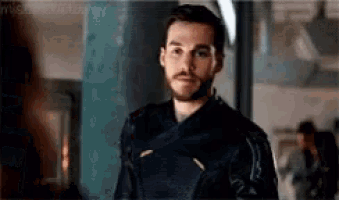 a man with a beard is wearing a black jacket and standing in front of a window .
