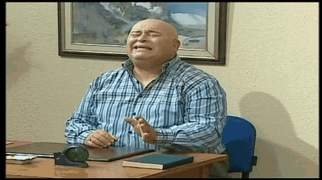 a bald man in a plaid shirt sits at a desk with his eyes closed