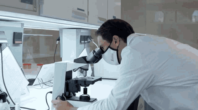 a man in a lab coat looking through a microscope with a mask on