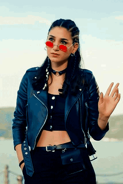 a woman wearing a leather jacket and red sunglasses is waving