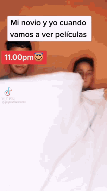a man and a woman are laying under a white blanket and a tiktok video shows them watching a movie .