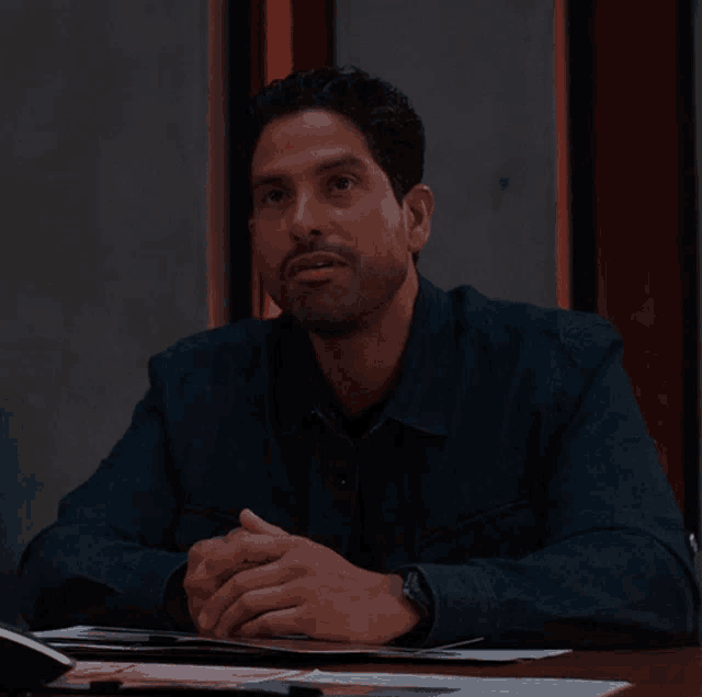a man in a blue shirt sits at a table with his hands folded