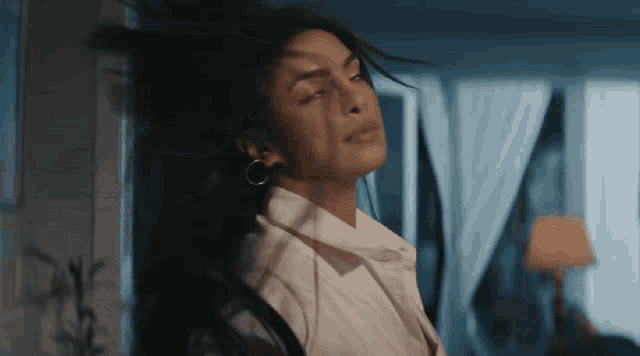 a woman in a white shirt is standing in front of a window and her hair is blowing in the wind
