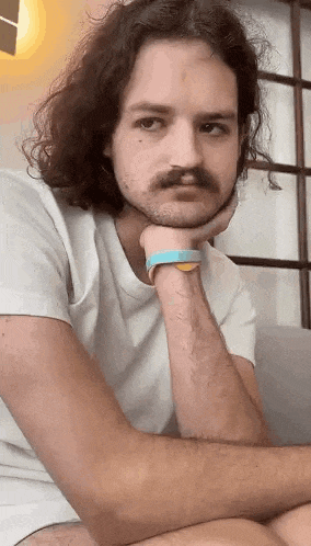 a man with long hair and a mustache is sitting on a couch with his hand on his chin and looking at the camera .