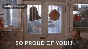 a man in a suit and tie is sitting in front of a door in a restaurant and saying `` so proud of you ! ''