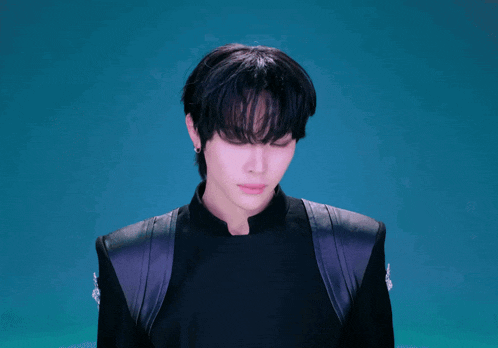 a young man with black hair and pink lips looks at the camera with a blue background
