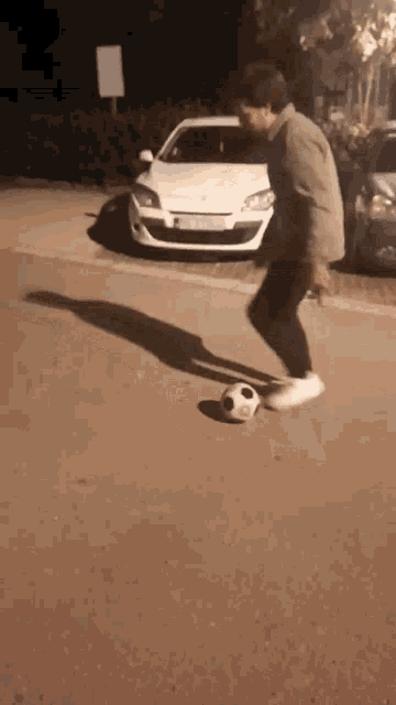 a man is kicking a soccer ball on the sidewalk in front of a white car