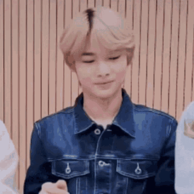 a young man with blonde hair is wearing a denim jacket and standing in front of a wooden wall .