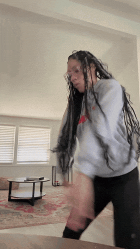 a woman with braids is dancing in a living room with a round coffee table in the background