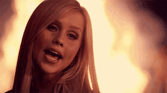 a woman with long blonde hair is standing in front of a fire