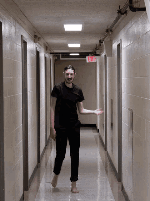 a man in a black shirt is walking down a hallway with an exit sign above him