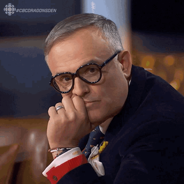 a man with glasses and a ring on his finger is sitting with his hand on his chin and the caption #cbcdragonsden