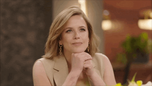 a woman wearing a tan jacket and gold earrings is sitting with her hands folded