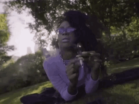 a woman is laying on the grass smoking a cigarette while wearing sunglasses .