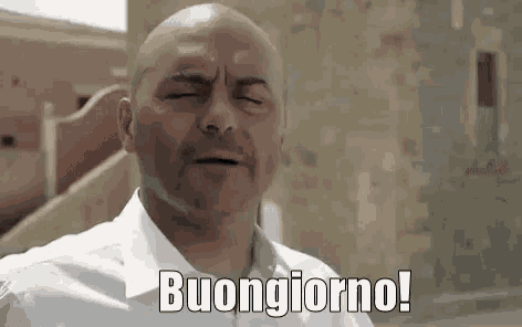 a bald man in a white shirt is standing in front of a building with his eyes closed and saying buongiorno .