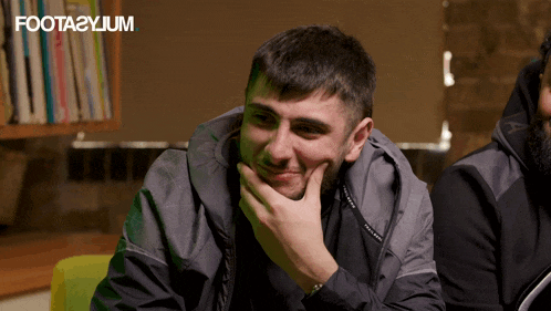 a man with his hand on his chin is smiling in front of a bookshelf that says foota2ylum