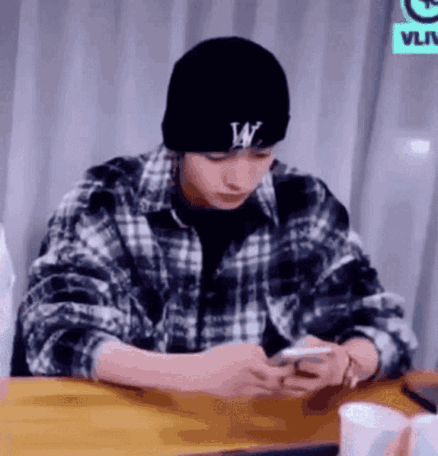 a man wearing a plaid shirt and a beanie is sitting at a table looking at his phone .