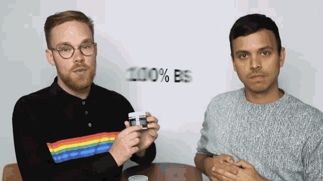 two men are sitting at a table and one of them is holding a bottle that says 100 % bs