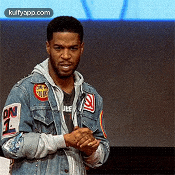 a man wearing a denim jacket with patches on the sleeves is standing in front of a blue wall .