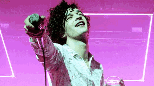 a man with curly hair singing into a microphone with a pink background