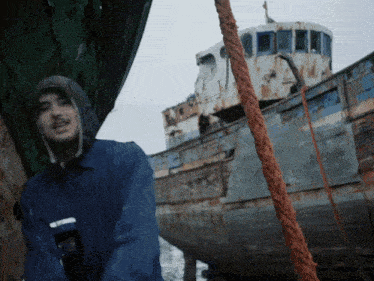 a man in a blue jacket stands in front of a rusty boat