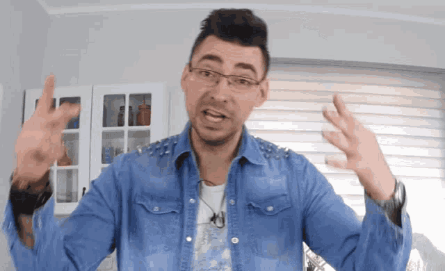 a man wearing glasses and a blue denim shirt is making a gesture with his hands