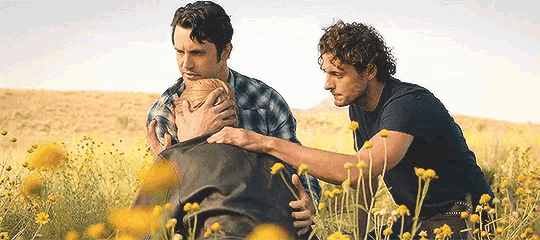 three men are sitting in a field of yellow flowers one of them is holding another man 's head
