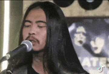 a man with long hair and a beard sings into a microphone in front of a sign that says ' ata '