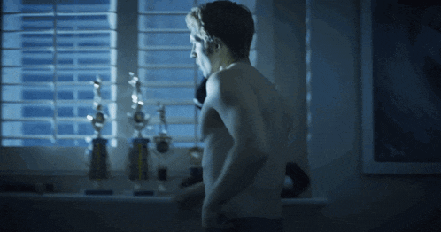 a man without a shirt is standing in front of a window with trophies on it