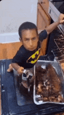 a little boy wearing a batman shirt is standing in front of a pan of food in an oven .