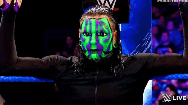 a wrestler with a green and purple face paint is standing in front of a crowd with his arms outstretched .