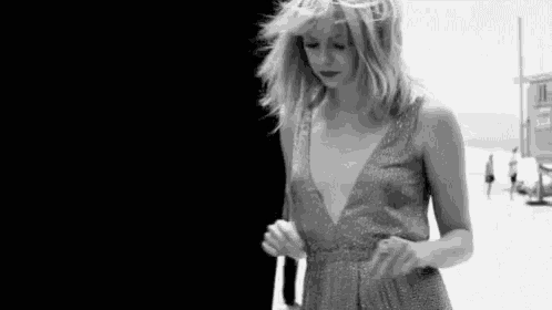 a black and white photo of a woman in a plunging neckline dress walking on a beach .