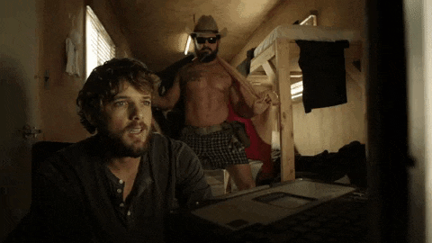 a man in a cowboy hat stands next to a man in underwear looking at a laptop