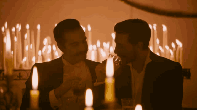 two men are sitting at a table with candles and one is drinking from a glass