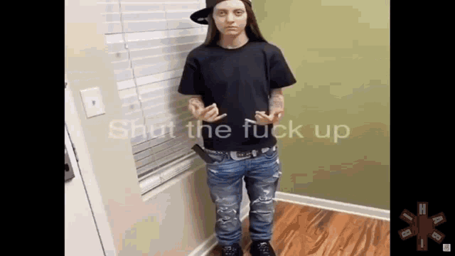 a woman in a black shirt and blue jeans is standing in front of a window with the words shut the fuck up on the bottom .