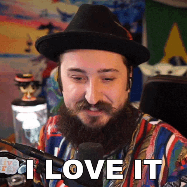 a man with a beard wearing a hat says " i love it " in front of a microphone