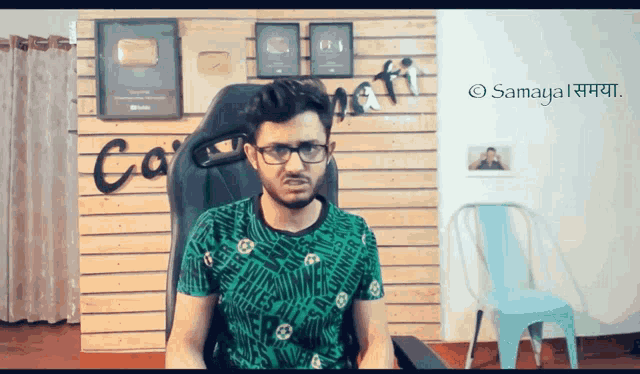 a man wearing glasses sits in a chair in front of a wall that has the word samaya on it